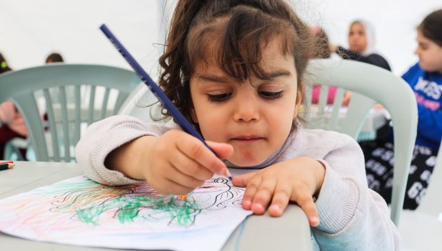 A young girl immersed in coloring a picture with crayons in a lively preschool setting.
